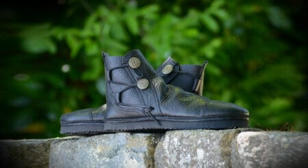 Black leather boots, reminiscent of custom moccasins with decorative buttons, rest elegantly on a stone surface, framed by a backdrop of blurred green foliage.