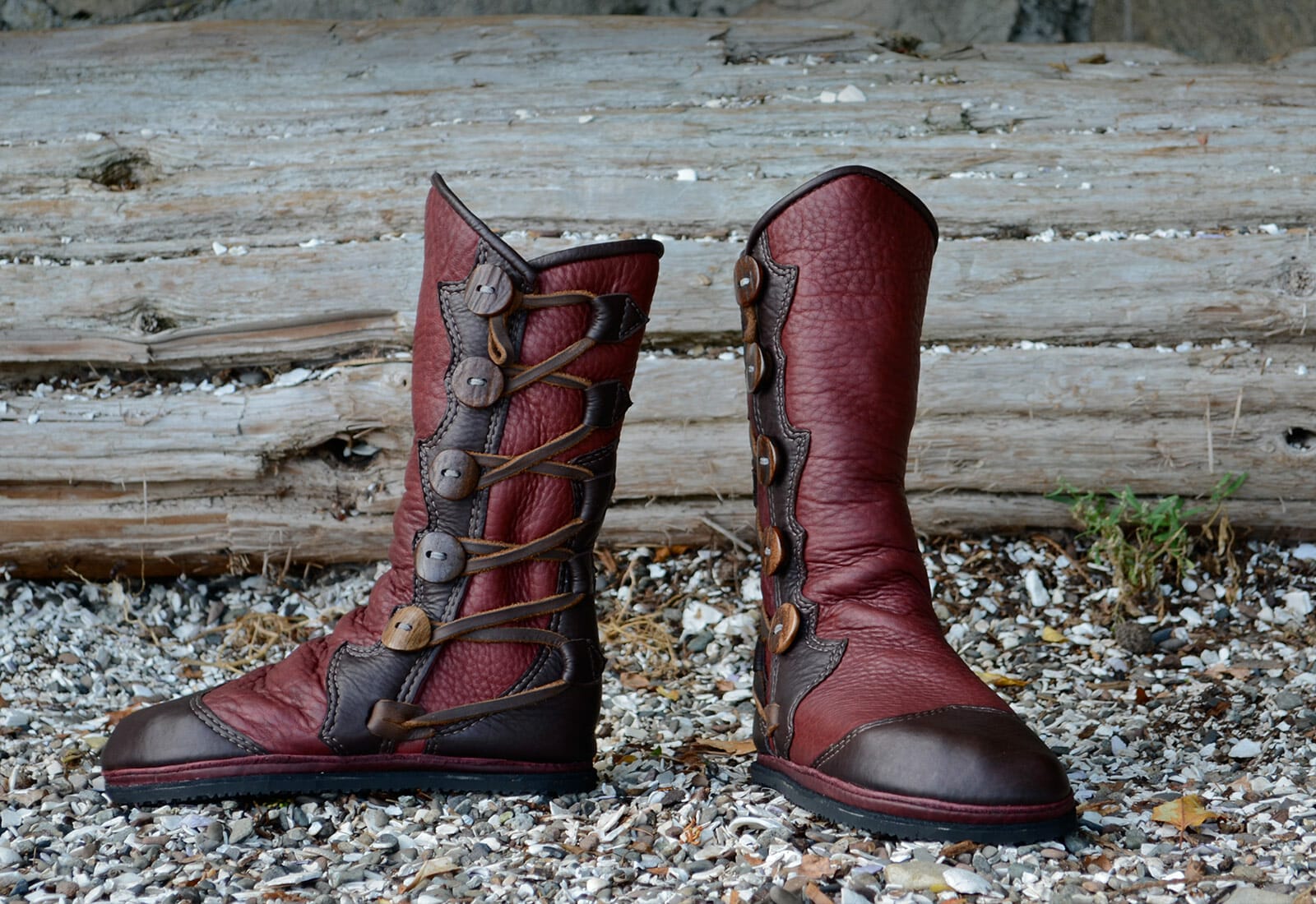 A pair of rustic red and brown leather boots, reminiscent of custom moccasins with side buttons, set against a background of wooden logs and scattered pebbles.