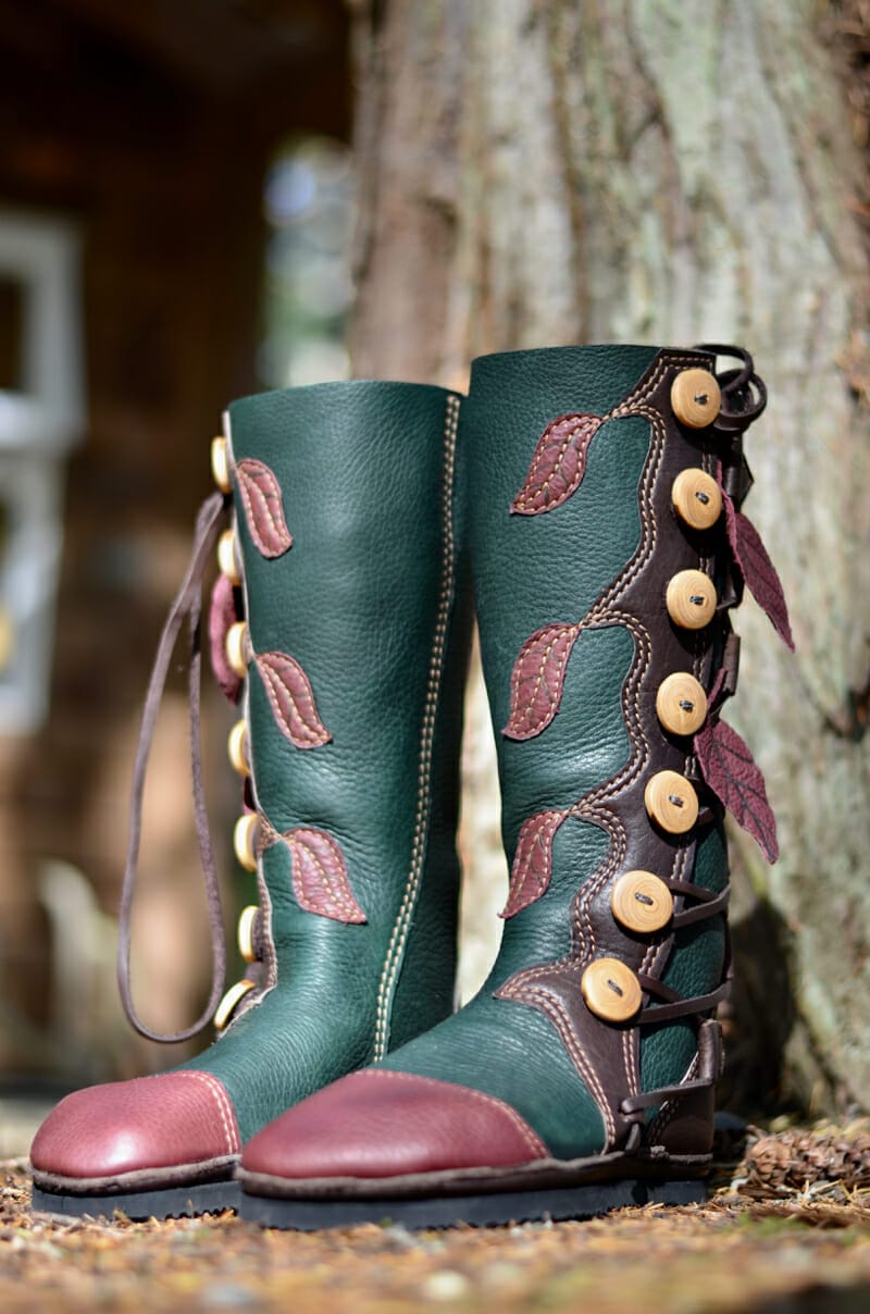 Green leather boots with maroon toes, decorated with leaf motifs and wooden buttons, offer custom style as they stand outdoors in front of a tree trunk.