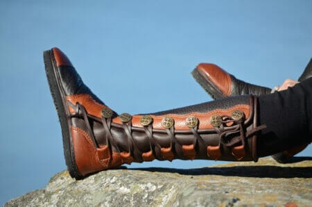 A custom pair of brown leather boots with ornate brass buttons and laces, stretched out on a rocky surface against a clear blue sky.