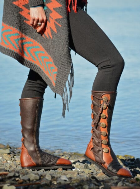 Person in black leggings and a patterned shawl stands on rocks near water, wearing high brown boots with side button details, offering a custom blend of style and function.