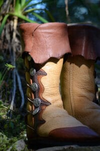Brown leather boots with decorative buttons stand on a forest floor, surrounded by the lush greenery of custom options in nature, as sunlight filters through the trees in the background.