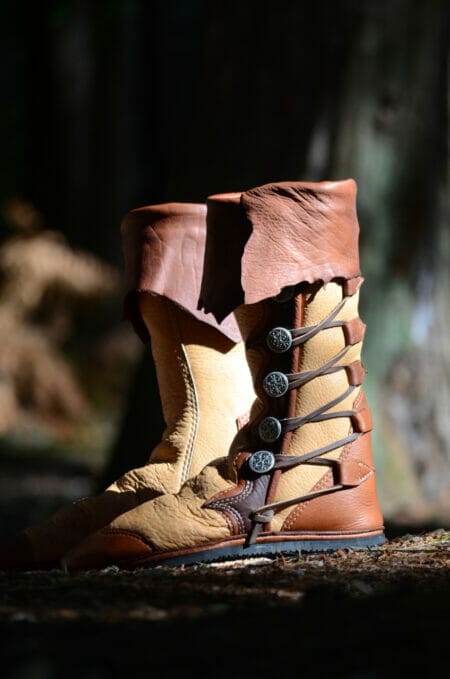 Leather boots with decorative lace-up sides and metal buttons, featuring a two-tone design in tan and brown, are set against a natural outdoor background. Discover custom options to tailor these boots to your style preferences.