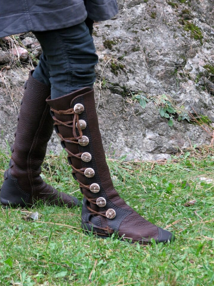 Person wearing tall, brown, leather boots with large round metal buttons and laces, standing on grass near a rocky surface—perfect for showcasing your style with our Custom Options Guide.